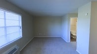 This single-story space presents a straightforward and functional layout designed for practical living. The main room is generously proportioned, featuring wall-to-wall carpet in a neutral tone that lends a sense of warmth underfoot. The walls are finished in a soft beige hue, creating a calm, understated backdrop that works well with a variety of furnishings and décor styles. A textured ceiling adds subtle visual interest overhead, while clean white baseboards frame the perimeter of the room, providing a crisp contrast to the neutral walls and flooring. Electrical outlets are conveniently positioned along the walls, and natural light filters in through a window on the left side of the room, brightening the interior. Visible through an open doorway in the far right corner, an adjoining bathroom is finished with matching neutral tones, a toilet, and a towel bar, indicating a complete and functional fixture arrangement. The transition between the carpeted main room and the hard-flooring bathroom is cleanly defined by white door trim. Overall, the space offers a blank-canvas quality, making it highly adaptable for a range of uses and personal styles.