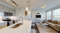 This single-floor residence showcases a thoughtfully designed open-concept layout that seamlessly connects the kitchen, dining, and living areas. The kitchen is a functional centerpiece, outfitted with gray cabinetry, stainless steel appliances including a refrigerator, electric range, and microwave, and a white quartz countertop that extends into a spacious island. The island accommodates bar seating with stylish patterned stools and is illuminated by a trio of pendant lights with brass fixtures, complemented by a track lighting system overhead. A marble-style backsplash adds a refined touch to the cooking area.

The open living room receives generous natural light through a bank of large windows dressed with sheer white curtains, offering outdoor views. Dark hardwood-style flooring runs throughout, grounding the bright, neutral palette. A wall-mounted media unit serves as the room's focal point.

The dining nook is a cozy, light-filled space positioned near a window, creating an inviting area for meals. The bedroom is comfortably proportioned with a window providing natural light and outdoor views.

The bathroom features a white quartz vanity with undermount sink, gray cabinetry, a large mirror with vanity lighting, and a full soaking tub with shower combination, finished with clean white tile.
