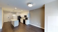 This single-floor space showcases an open-concept layout that seamlessly connects the kitchen and living areas. The kitchen is well-appointed with white shaker-style cabinetry, a light-toned stone countertop island with pendant lighting overhead, and black appliances including a refrigerator, range, and over-the-range microwave. A stainless steel sink is positioned along the back counter, complemented by recessed lighting that brightens the workspace. The living area flows naturally from the kitchen, featuring warm, dark hardwood-style flooring that runs throughout the space, creating a cohesive and grounded aesthetic. Neutral taupe-toned walls provide a versatile backdrop, while white baseboards offer a clean contrast. Ceiling-mounted lighting in the living area ensures consistent illumination. The overall design balances functional layout with contemporary finishes, offering a practical yet polished environment. The open sightlines between the kitchen and adjoining space enhance the sense of roominess, making the layout feel connected and airy.