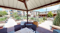 This inviting outdoor amenity space centers around a covered pavilion with a pitched roof supported by warm wood-toned beams, offering a shaded retreat that opens directly to a lush community garden. The pavilion is furnished with wicker lounge chairs, cushioned seating, and a wooden dining table, creating a versatile area for relaxation and gathering. A curved brick knee wall lines the perimeter of the pavilion, adding a classic architectural touch that seamlessly transitions to the surrounding hardscape.

Extending from the pavilion, a wide concrete pathway leads through an expansive community garden area featuring numerous raised wooden planter beds filled with a diverse mix of flowering plants, vegetables, and ornamental grasses. The raised beds are thoughtfully arranged on either side of the walkway, encouraging an immersive garden experience. Timber-style benches and cylindrical wooden waste receptacles are strategically placed throughout, enhancing both functionality and aesthetic appeal.

The landscape is enriched with mature ornamental shrubs and trees that provide visual interest and natural privacy. The open-sky setting allows abundant natural light to fill the space, and views of the surrounding multi-story residential buildings frame the garden, reinforcing the sense of a well-integrated community environment. The overall ambiance is fresh, green, and welcoming.