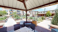 This inviting outdoor amenity space centers around a covered pavilion with a pitched roof supported by warm wood-toned beams, offering a shaded retreat that opens directly to a lush community garden. The pavilion is furnished with wicker lounge chairs, cushioned seating, and a wooden dining table, creating a versatile area for relaxation and gathering. A curved brick knee wall lines the perimeter of the pavilion, adding a classic architectural touch that seamlessly transitions to the surrounding hardscape.

Extending from the pavilion, a wide concrete pathway leads through an expansive community garden area featuring numerous raised wooden planter beds filled with a diverse mix of flowering plants, vegetables, and ornamental grasses. The raised beds are thoughtfully arranged on either side of the walkway, encouraging an immersive garden experience. Timber-style benches and cylindrical wooden waste receptacles are strategically placed throughout, enhancing both functionality and aesthetic appeal.

The landscape is enriched with mature ornamental shrubs and trees that provide visual interest and natural privacy. The open-sky setting allows abundant natural light to fill the space, and views of the surrounding multi-story residential buildings frame the garden, reinforcing the sense of a well-integrated community environment. The overall ambiance is fresh, green, and welcoming.