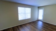 This single-floor space offers a clean, functional layout with notable interior finishes. The main living area showcases rich, dark hardwood flooring that contrasts attractively with the neutral beige walls, creating a warm yet understated aesthetic. A textured popcorn ceiling adds dimension overhead while maintaining a classic residential character.

Natural light is a defining feature of this space, provided by a large, multi-pane window fitted with horizontal blinds, which allows for adjustable light control. The window offers views of surrounding greenery, bringing a sense of the outdoors inside. Adjacent to the window, a full-length exterior door with a frosted glass panel further enhances brightness and provides direct outdoor access.

The room is well-proportioned, offering comfortable open space suitable for a variety of furniture arrangements. White baseboards frame the perimeter, adding a clean, finished look to the flooring transition. The overall ambiance is bright, neutral, and move-in ready, making it a versatile space adaptable to various interior styles and personal preferences.