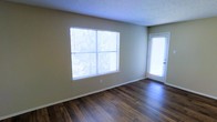 This single-floor space offers a clean, functional layout with notable interior finishes. The main living area showcases rich, dark hardwood flooring that contrasts attractively with the neutral beige walls, creating a warm yet understated aesthetic. A textured popcorn ceiling adds dimension overhead while maintaining a classic residential character.

Natural light is a defining feature of this space, provided by a large, multi-pane window fitted with horizontal blinds, which allows for adjustable light control. The window offers views of surrounding greenery, bringing a sense of the outdoors inside. Adjacent to the window, a full-length exterior door with a frosted glass panel further enhances brightness and provides direct outdoor access.

The room is well-proportioned, offering comfortable open space suitable for a variety of furniture arrangements. White baseboards frame the perimeter, adding a clean, finished look to the flooring transition. The overall ambiance is bright, neutral, and move-in ready, making it a versatile space adaptable to various interior styles and personal preferences.