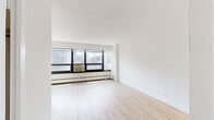Located at 3130 N Lake Shore Drive in Chicago's Lakeview neighborhood, this unit offers a bright and clean living environment with a minimalist aesthetic throughout. The main living area is generously proportioned, featuring light-toned hardwood flooring and crisp white walls that create an airy, open atmosphere. A wide bank of floor-to-ceiling windows with black frames floods the space with natural light and frames expansive views of the surrounding Chicago cityscape, making it a standout feature of the living room. A baseboard heating system runs beneath the windows, complemented by a wall-mounted air conditioning unit for year-round climate control. The layout connects seamlessly to a separate bedroom area, also finished with light wood flooring and bright white walls, providing a calm and neutral retreat. The kitchen is efficiently designed with white cabinetry, dark granite-style countertops, a stainless steel sink, a coil-top range, and a full-size refrigerator, all finished with light tile flooring. The open flow between the living space, bedroom, and kitchen makes the unit highly functional. Overall, the space maintains a fresh, well-maintained appearance with consistent finishes throughout.
