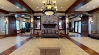 This single-floor space exudes a refined, traditional aesthetic characterized by an impressive blend of architectural craftsmanship and warm, inviting design elements. The central lobby serves as the focal point, anchored by a dramatic floor-to-ceiling stacked-stone fireplace flanked by a carved wood mantel and decorative shelving. An ornate wrought-iron chandelier with frosted glass shades hangs overhead, complemented by recessed lighting and wall sconces that collectively create a layered, warm ambiance.

Dark wood ceiling beams and wide baseboards frame the space throughout, providing rich contrast against the neutral beige tile flooring, which features inlaid wood-toned tile strips for added visual interest. Traditional seating arrangements — including carved wooden sofas and armchairs upholstered in muted earth tones — are thoughtfully positioned across multiple seating areas.

Arched doorways with decorative columns and textured pilasters lead into secondary areas, where additional seating nooks benefit from natural light streaming through dark wood-trimmed windows, offering views of palm trees outside. A work or reception area with classic wooden furniture is also present. Throughout, wall sconces, decorative accents, and lush greenery contribute to a cohesive, upscale traditional character.