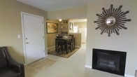 This single-floor residence offers a warm and inviting atmosphere, thoughtfully designed for comfortable everyday living. The living area serves as the heart of the home, anchored by a classic fireplace with a sleek dark surround — an ideal focal point for relaxing gatherings. Soft, neutral-toned carpeting flows throughout, complementing the warm beige walls that lend the space a cozy, understated elegance. A decorative starburst wall accent adds a stylish touch to the living area. The open layout connects seamlessly to the dining space, which comfortably accommodates a dark-wood dining table and chair set, set atop a patterned area rug. Built-in shelving along the dining area wall provides both display and storage opportunities, enhancing the functional character of the space. A framed landscape artwork adds a curated, homey feel. The kitchen is conveniently accessible from the dining area and features white appliances and ample cabinetry. Tile flooring near the entryway transitions smoothly into the carpeted living spaces, defining the entry zone while maintaining a cohesive flow throughout the home. Overall, this residence balances practicality with comfort in a well-connected, single-level layout.