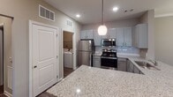 Located at 6038 Hixson Pike in Hixson, Tennessee, this single-floor residence offers a thoughtfully designed open-concept layout with a neutral color palette and consistent finishes throughout. The kitchen is a functional centerpiece, featuring gray shaker-style cabinetry, a white subway tile backsplash, granite countertops, a large island with pendant lighting, and a full suite of stainless steel appliances including a refrigerator, range, and microwave. An adjacent in-unit laundry closet houses a full-size top-load washer and dryer with overhead shelving. The open living area is generously proportioned, finished with plush wall-to-wall carpeting, crown molding, and a ceiling fan. Natural light filters in through windows fitted with horizontal blinds, lending the space a bright, comfortable feel. Additional rooms provide flexible spaces for bedrooms or office use, each carpeted and equipped with ceiling fans. The bathrooms feature granite-topped vanities with gray cabinetry, hardwood-style flooring, and tub-shower combinations with brushed nickel fixtures. Clean architectural lines, consistent warm-toned walls, and quality finishes contribute to a cohesive, move-in-ready living environment.