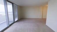This single-floor residence offers a clean, open layout designed for comfortable everyday living. The main living area is generously proportioned, featuring wall-to-wall neutral carpeting that lends a warm and understated tone throughout. A striking floor-to-ceiling window fitted with horizontal blinds dominates one side of the room, flooding the interior with natural light while offering the option for privacy and light control. The walls are finished in a soft, creamy white tone that enhances the sense of brightness and openness. The room's layout is notably flexible, providing ample space for a variety of furniture arrangements to suit different living needs. Subtle architectural detailing, including a gentle step in the wall plane toward the far end of the room, adds a quiet visual interest to the otherwise minimalist design. The space is vacant, making it easy to envision and personalize to one's own style. Overall, the living area presents a calm, neutral ambiance with a functional and adaptable floor plan, well-suited for those seeking a bright and open interior environment.