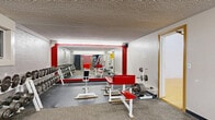 Located at 1800 Fifth Ave NE in East Grand Forks, Minnesota, this fitness center offers a comprehensive, multi-zone workout facility spread across a generously sized single floor. The weight room serves as a central hub, featuring an extensive dumbbell rack with a full range of weights, multiple benches with red and white upholstery, barbells, plate storage, and squat racks. Bold red accent columns and large wall mirrors enhance the functional, energetic atmosphere of this area. Fluorescent lighting throughout ensures a well-lit environment for all activities. An adjoining machine room runs the length of the facility, lined with a broad selection of Life Fitness and similarly branded cable and resistance machines, providing a complete circuit-training experience. A staircase connects areas within the facility, adding a sense of depth and flow to the layout. The racquetball court stands out with its classic golden hardwood floor, high-reaching white walls, and green court markings — accessible directly from the weight area via a connecting door with a pass-through window. A dedicated tanning room, finished in warm yellow tones, houses a Montego Bay tanning bed and includes a wooden bench and wall-mounted sanitation supplies, offering a relaxing complement to the workout amenities.