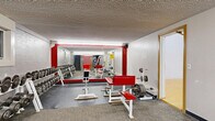 Located at 1800 Fifth Ave NE in East Grand Forks, Minnesota, this fitness center offers a comprehensive, multi-zone workout facility spread across a generously sized single floor. The weight room serves as a central hub, featuring an extensive dumbbell rack with a full range of weights, multiple benches with red and white upholstery, barbells, plate storage, and squat racks. Bold red accent columns and large wall mirrors enhance the functional, energetic atmosphere of this area. Fluorescent lighting throughout ensures a well-lit environment for all activities. An adjoining machine room runs the length of the facility, lined with a broad selection of Life Fitness and similarly branded cable and resistance machines, providing a complete circuit-training experience. A staircase connects areas within the facility, adding a sense of depth and flow to the layout. The racquetball court stands out with its classic golden hardwood floor, high-reaching white walls, and green court markings — accessible directly from the weight area via a connecting door with a pass-through window. A dedicated tanning room, finished in warm yellow tones, houses a Montego Bay tanning bed and includes a wooden bench and wall-mounted sanitation supplies, offering a relaxing complement to the workout amenities.