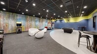 Located at 50 Morgan St in Hartford, Connecticut, this dynamic game room delivers a generously sized, single-floor entertainment space designed for recreation and social engagement. The interior is defined by a bold two-tone color scheme — deep navy blue on the lower walls transitioning to a vivid chartreuse yellow near the ceiling — creating a lively and energetic atmosphere throughout.

The space features a distinctive curved floor design, where a smooth white surface flows organically into carpeted sections, adding visual interest and helping to define different activity zones. A dark drop-tile ceiling with recessed lighting provides even illumination across the room.

A standout artistic element is the large-scale abstract mural that spans an entire wall, featuring a rich mix of colors that adds a gallery-like dimension to the space. Recreational amenities are plentiful and well-distributed: a full-size pool table anchors one zone, a backlit air hockey table commands attention along the mural wall, and a row of classic pinball machines and a Pac-Man arcade cabinet line the opposite side.

The lounge area is furnished with sculptural, modern white seating — including a curved sofa and rounded pod chairs — paired with a low-profile wooden coffee table, offering a comfortable retreat between games. High-top tables and bar chairs provide additional seating options, making this space equally suited for casual gatherings and competitive play.