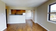 This single-floor residence offers a thoughtfully designed open-concept layout that seamlessly connects the living, dining, and kitchen areas into one cohesive living space. Rich, warm-toned hardwood flooring runs throughout, lending a sense of continuity and warmth to the entire floor plan.

The kitchen is positioned at the rear of the open layout and features classic honey-toned wood cabinetry, a mosaic tile backsplash, and stainless steel appliances. A raised half-wall separates the kitchen from the living area, providing both functional counter space and a natural visual boundary without compromising the open feel of the space.

The living and dining areas are generously proportioned, offering ample room for a variety of furniture arrangements. A window along the side wall allows natural light to filter in, brightening the neutral beige walls and contributing to the overall airy ambiance. A wall-mounted heating unit sits beneath the window, providing a practical heating solution.

The textured ceiling adds a subtle architectural detail that complements the overall design. Clean white baseboards trim the perimeter of the space, providing a crisp contrast against both the flooring and the walls. The overall atmosphere is warm, practical, and move-in ready, suited for comfortable everyday living.