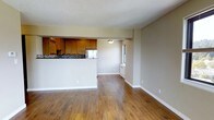 This single-floor residence offers a thoughtfully designed open-concept layout that seamlessly connects the living, dining, and kitchen areas into one cohesive living space. Rich, warm-toned hardwood flooring runs throughout, lending a sense of continuity and warmth to the entire floor plan.

The kitchen is positioned at the rear of the open layout and features classic honey-toned wood cabinetry, a mosaic tile backsplash, and stainless steel appliances. A raised half-wall separates the kitchen from the living area, providing both functional counter space and a natural visual boundary without compromising the open feel of the space.

The living and dining areas are generously proportioned, offering ample room for a variety of furniture arrangements. A window along the side wall allows natural light to filter in, brightening the neutral beige walls and contributing to the overall airy ambiance. A wall-mounted heating unit sits beneath the window, providing a practical heating solution.

The textured ceiling adds a subtle architectural detail that complements the overall design. Clean white baseboards trim the perimeter of the space, providing a crisp contrast against both the flooring and the walls. The overall atmosphere is warm, practical, and move-in ready, suited for comfortable everyday living.