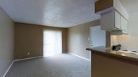 This single-floor space features an open living area finished with light-colored wall-to-wall carpeting that contributes to a clean, understated aesthetic. The room is characterized by its textured popcorn ceiling, which adds a classic touch to the interior. A large sliding glass door, dressed with vertical blinds, serves as the primary source of natural light, brightening the living space and providing access to the outdoors. The warm beige-toned walls create a neutral backdrop throughout the room. A standard entry door is also visible, positioned along the interior wall. Adjacent to the living area, a partial view of the kitchen reveals a dark-toned countertop, suggesting a defined separation between the living and kitchen spaces. The layout appears straightforward and practical, with an open flow between the kitchen and living room. White baseboards provide a clean contrast against the carpeted floors and walls. Overall, the space presents a simple, functional design suited for everyday living.