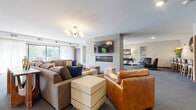 This single-floor residential community offers a refined selection of shared spaces designed for comfort, social engagement, and everyday convenience. The central lounge serves as the heart of the community, featuring a striking stone-clad fireplace column with a wall-mounted television, a built-in linear gas fireplace, and a warm mix of plush sectional seating, a cognac leather armchair, and a geometric wood ottoman — all bathed in natural light from large windows dressed in sheer drapery. A sputnik-style brass chandelier adds a contemporary finishing touch overhead. Adjacent gathering areas extend the social spaces, offering lounge seating arrangements with modern accent furniture, curated wall art, and a high-top bar counter ideal for casual socializing. A dedicated dining room accommodates a large group around a glass-topped table surrounded by upholstered chairs, with views to the outdoors through generous windows. A kitchenette room provides a full-size refrigerator, mosaic tile backsplash, and bar-height seating for informal dining or entertaining. The wide, carpeted corridors are appointed with decorative wall art, sculptural accents, and warm sconce lighting, creating a welcoming passage throughout the building. A mezzanine-style seating nook with a wrought-iron railing and pendant chandelier completes the community's thoughtfully curated design.