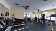 This fitness center offers a comprehensive workout environment within a single-level layout. The space is finished with dark rubberized flooring, providing both durability and traction throughout the exercise area. Warm yellow-beige walls with dark wainscoting lend a structured, clean aesthetic to the room.

Cardio equipment, including treadmills and an elliptical machine, lines one side of the room, while a dedicated strength training area features a cable machine, weight rack, and multi-station resistance equipment on the other. The open floor plan allows for comfortable movement between stations.

Natural light streams in through multiple windows fitted with horizontal blinds along the far wall, creating a bright and energizing atmosphere. Two ceiling fans with light fixtures are mounted overhead, promoting air circulation throughout the space. Recessed lighting supplements the natural light for consistent illumination. A mirrored wall section near the back of the room enhances the sense of depth and allows users to monitor their form during workouts. The overall layout is functional and thoughtfully organized for a varied fitness routine.