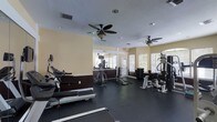 This fitness center offers a comprehensive workout environment within a single-level layout. The space is finished with dark rubberized flooring, providing both durability and traction throughout the exercise area. Warm yellow-beige walls with dark wainscoting lend a structured, clean aesthetic to the room.

Cardio equipment, including treadmills and an elliptical machine, lines one side of the room, while a dedicated strength training area features a cable machine, weight rack, and multi-station resistance equipment on the other. The open floor plan allows for comfortable movement between stations.

Natural light streams in through multiple windows fitted with horizontal blinds along the far wall, creating a bright and energizing atmosphere. Two ceiling fans with light fixtures are mounted overhead, promoting air circulation throughout the space. Recessed lighting supplements the natural light for consistent illumination. A mirrored wall section near the back of the room enhances the sense of depth and allows users to monitor their form during workouts. The overall layout is functional and thoughtfully organized for a varied fitness routine.