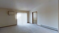 This single-floor space presents a clean and functional layout with a focus on simplicity. The main room is finished with wall-to-wall light-toned carpeting that contributes to a warm, quiet atmosphere. A large window dressed with vertical blinds spans a significant portion of one wall, allowing an abundance of natural light to fill the room while offering privacy control. The textured popcorn ceiling adds a classic, understated character to the space. A baseboard heater runs along the far wall, providing an efficient and unobtrusive heating solution. Neutral-toned walls with warm wood trim framing the doorways and baseboards create a cohesive, traditional aesthetic throughout. An open doorway connects the main room to an adjacent space, suggesting a straightforward and accessible floor plan. The overall ambiance is bright, airy, and move-in ready, offering a blank canvas for personalization. The space is well-suited for those seeking a no-frills, comfortable living environment with practical features and ample natural light.