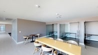 This thoughtfully designed single-floor space offers a refined blend of communal and private areas suited for work, meetings, or relaxation. The open lounge and dining area anchors the space with a large light wood communal table surrounded by contemporary curved chairs, complemented by smaller bistro-style tables for more intimate seating arrangements. Light-toned wood-effect flooring runs throughout, contributing to a clean, cohesive aesthetic.

A standout architectural feature is the set of fully glazed, floor-to-ceiling glass partition rooms with sleek metal hardware. These enclosed spaces provide privacy without sacrificing visual openness, each furnished with minimalist desks and benches — ideal for focused work or small meetings. Natural light filters in through large windows, brightening the main lounge area and offering views to the surrounding streetscape.

The walls are painted in soft, muted tones — warm greige in the main area and a calming light blue in the lounge — adorned with curated contemporary artwork, including a triptych of bold, colorful prints and a paired set of abstract teal canvases. Recessed lighting and white ceilings enhance the bright, airy atmosphere throughout. The overall ambiance is professional yet welcoming, with modern finishes and thoughtful design details creating a versatile and comfortable environment.
