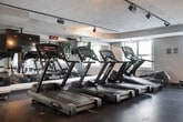 Located at 1100 Avalon Square in Glen Cove, NY, this well-equipped fitness center offers a comprehensive workout environment on a single floor. The space features durable dark rubber flooring throughout, providing a practical and professional surface suited for a wide range of exercise activities. Track lighting runs along the textured ceiling, ensuring the entire space is well-illuminated, while multiple windows allow natural light to filter in, creating a bright and welcoming atmosphere. Large wall-mounted mirrors line one side of the room, visually expanding the space and allowing users to monitor their form during workouts. The cardio section is outfitted with multiple treadmills, an elliptical trainer, a stationary bike, and a rowing machine, catering to a variety of cardiovascular training preferences. A multi-functional cable and weight machine serves as the centerpiece for strength training, complemented by a dedicated free weights area featuring a full dumbbell rack. A wall-mounted flat-screen television adds an entertainment element to the space. A separate interior window and access door suggest an adjoining office or attendant area, adding a layer of convenience and oversight to the facility. Overall, the fitness center delivers a clean, functional, and well-appointed environment for residents seeking a convenient on-site workout facility.