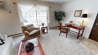 This single-floor residence offers a thoughtfully arranged layout that balances comfort and functionality across several distinct living areas. The living room serves as the heart of the home, highlighted by a large multi-pane window dressed with flowing white drapes that invite generous natural light and offer views of the surrounding greenery. Plush wall-to-wall carpeting, baseboard heating, and a wall-mounted air conditioning unit contribute to year-round comfort. A glass-topped coffee table and upholstered seating add to the relaxed atmosphere, while a dedicated desk area provides a functional workspace.

The dining area features a round glass table with tufted leather chairs beneath a ceiling fan with light fixture, creating an intimate setting for meals. The kitchen is efficiently arranged with rich cherry-toned cabinetry, a laminate countertop, stainless steel sink, and white appliances including a refrigerator and dishwasher.

Two carpeted bedrooms offer restful retreats; one presents a spacious sliding-door closet, while the other carries a soft, light-filled aesthetic with neutral tones. The full bathroom includes a tub-shower combination, a wide vanity mirror, and tile surround, finished in a clean, neutral palette throughout.