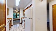 This single-floor unit offers a practical and straightforward layout connecting a kitchen, living area, dining space, bedroom, and bathroom. The kitchen is equipped with warm honey-toned wood cabinetry, light-colored countertops, a white electric range with a ventilation hood, a full-size refrigerator, and tile flooring. A skylight-style ceiling fixture provides overhead illumination, while a pass-through view connects the kitchen to the adjacent carpeted dining area, which features a ceiling fan and a window that brings in natural light from outside.

The living room is a generously proportioned, open space with neutral carpet, white walls, and a distinctive brick column accent that adds architectural character. A baseboard heating unit runs along one wall, providing climate control throughout the room. The bedroom is a quiet, carpeted room with neutral tones and a solid wood door, offering a clean and simple space.

The bathroom features a full tub and shower combination with chrome fixtures and a clean white surround, complemented by tile flooring. Overall, the unit presents a functional and cohesive interior with consistent warm wood accents, neutral finishes, and practical amenities throughout.
