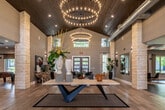 This single-floor space presents a bold and sophisticated design that seamlessly blends contemporary style with rustic architectural elements. The grand entrance lobby immediately captivates with its soaring vaulted ceiling clad in rich wood planking, flanked by prominent limestone columns and anchored by two dramatic tiered ring chandeliers that cast a warm, inviting glow over the light hardwood floors below.

From the entry, the space opens into a series of distinct yet connected zones. A generously proportioned lounge area features a large sectional sofa, a wall-mounted fireplace, a flat-screen television, and curated accent furniture, creating a comfortable and stylish gathering hub. Adjacent to this is a wet bar and counter seating area illuminated by sculptural pendant lighting, ideal for social gatherings.

A dedicated workspace or leasing area is defined by bold orange accent walls, a vivid yellow patterned rug, sleek pendant lighting, and colorful wall art, lending an energetic and creative atmosphere. Nearby, a herringbone-tiled foyer with a teal accent niche and vibrant abstract artwork creates a refined transition between areas.

Throughout the space, exposed ductwork, clerestory windows, and large glass entry doors contribute to an open, airy industrial-contemporary aesthetic, while curated art, greenery, and eclectic furnishings add warmth and personality to every corner.