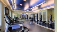 Located at 8110 Six Forks Rd in Raleigh, NC, this fitness center offers a comprehensive and thoughtfully designed workout environment on a single floor. The space is immediately striking, featuring a dramatic ceiling finished in deep blue tones with recessed and pendant lighting that creates an energizing yet polished atmosphere. Large wall-to-wall mirrors line one side of the facility, visually expanding the space and allowing users to monitor their form throughout their workouts. The flooring transitions between light and dark zones, subtly delineating cardio and strength training areas. The cardio section is well-equipped with multiple treadmills and other aerobic machines positioned for easy access. The strength training area features a comprehensive dumbbell rack with a wide range of free weights, along with multiple cable and resistance machines designed to target various muscle groups. A large flat-screen TV is mounted prominently on the mirrored wall, providing entertainment during workouts. The walls are finished in a warm yellow tone with louvered accents, lending a classic, upscale character to the space. The overall ambiance is clean, spacious, and professional, making it a well-appointed facility suited for a full range of fitness routines.