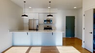 Located at 7960 SE Luther Rd in Portland, Oregon, this single-floor residence offers a contemporary design with clean lines and modern finishes throughout. The open-concept living and kitchen area serves as the heart of the home, anchored by a large central island with a white quartz countertop, an undermount sink, and seating overhang. Pendant lights with matte black fixtures hang above, complementing the white shaker-style cabinetry, textured tile backsplash, and full suite of stainless steel appliances including a French door refrigerator, electric range, and over-the-range microwave. Light hardwood flooring flows throughout, and large windows allow generous natural light into the living area, which opens to a private outdoor patio with views of the surrounding community. The bedrooms are bright and well-proportioned, with one offering direct patio access through sliding glass doors. The bathrooms are finished with navy blue shaker vanities, white quartz countertops, large mirrors, and stylish sconce lighting. One bathroom features a double-sink vanity, while another includes a full tub-shower combo. A dedicated closet houses a stacked washer and dryer, adding practical convenience to this well-appointed home.
