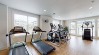 Located in Vancouver, Washington, this single-floor fitness center is a comprehensively outfitted exercise facility designed to support a wide range of workout routines. The space features warm hardwood-style flooring throughout, recessed lighting, and neutral-toned walls that create a clean, energizing environment. Large windows with horizontal blinds line multiple walls, allowing abundant natural light to fill the room while offering views of the surrounding neighborhood.

The cardio zone is generously stocked with multiple treadmills and elliptical trainers, providing ample options for aerobic exercise. A dedicated strength and free-weight area includes a multi-tiered dumbbell rack with a full range of weights, adjustable workout benches, and a wall-mounted flat-screen television for entertainment during training sessions. Full-length mirrors along one wall visually expand the space and allow users to monitor their form.

A standout feature is a large, dramatic wall mural depicting silhouetted figures atop a cliff at sunset, adding an inspiring, motivational focal point to the room. Additional equipment includes a cable machine, stationary bikes, and a stair climber. Exercise accessories such as stability balls, resistance tools, and yoga mats round out the offerings. Wall-mounted fans provide supplemental airflow, and a glass door offers direct access to an outdoor area, enhancing ventilation and convenience.