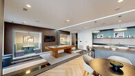 Located at 3015 Routh St in Dallas, TX, this single-floor amenity lounge is designed with a contemporary aesthetic that blends functionality and style. The open layout integrates several distinct zones, each thoughtfully furnished to encourage both social interaction and relaxed leisure.

At the heart of the space sits a substantial pool table anchored on a mixed-material floor featuring herringbone hardwood and inlaid tile sections, adding visual depth and defining different activity areas. Dark wood wall paneling provides a warm contrast to the crisp white ceilings and walls, while recessed lighting and linear LED cove accents illuminate the space evenly throughout.

A wet bar area features dark granite countertops, white cabinetry, a patterned tile backsplash, stainless steel appliances, and open shelving set against a striking teal accent wall. Adjacent to the bar, a dining table with pendant lighting offers a casual gathering spot. A cozy lounge nook with built-in seating and a wall-mounted television provides a more intimate setting.

The lounge seating area features richly upholstered furniture in warm amber and deep green tones. Large floor-to-ceiling glass doors flood the interior with natural light and open directly to an outdoor patio, seamlessly connecting the indoor and outdoor environments.