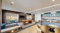 Located at 3015 Routh St in Dallas, TX, this single-floor amenity lounge is designed with a contemporary aesthetic that blends functionality and style. The open layout integrates several distinct zones, each thoughtfully furnished to encourage both social interaction and relaxed leisure.

At the heart of the space sits a substantial pool table anchored on a mixed-material floor featuring herringbone hardwood and inlaid tile sections, adding visual depth and defining different activity areas. Dark wood wall paneling provides a warm contrast to the crisp white ceilings and walls, while recessed lighting and linear LED cove accents illuminate the space evenly throughout.

A wet bar area features dark granite countertops, white cabinetry, a patterned tile backsplash, stainless steel appliances, and open shelving set against a striking teal accent wall. Adjacent to the bar, a dining table with pendant lighting offers a casual gathering spot. A cozy lounge nook with built-in seating and a wall-mounted television provides a more intimate setting.

The lounge seating area features richly upholstered furniture in warm amber and deep green tones. Large floor-to-ceiling glass doors flood the interior with natural light and open directly to an outdoor patio, seamlessly connecting the indoor and outdoor environments.