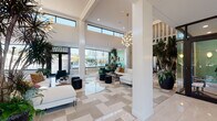 Located along River Road in North Little Rock, Arkansas, this single-floor clubhouse is a sophisticated amenity space designed with contemporary flair and an abundance of natural light. The grand entrance welcomes visitors with soaring double-height ceilings, polished large-format tile flooring in a neutral checkered pattern, and floor-to-ceiling windows that flood the interior with daylight and offer views of the surrounding landscape.

The main lounge area is expansive and elegantly appointed, featuring white sofas, black leather ottomans, emerald velvet accent chairs, and statement pendant lighting. A dramatic living green wall serves as a striking botanical focal point near the entryway, complemented by oversized tropical potted plants throughout the space. Vertical slatted wood screens define zones while maintaining an open, airy feel.

A central lounge zone showcases a suspended circular fireplace beneath a bold slatted wood ceiling medallion, creating an intimate gathering atmosphere. Adjacent to this is a full kitchen with white cabinetry and quartz countertops. The game area features a full-size billiards table set against large windows with views of the outdoor pool area. A bar-height dining nook and multiple seating clusters complete this dynamic, multi-functional clubhouse.