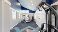 Located at 793 Patriot Pkwy in Rock Hill, SC, this dedicated exercise room offers a well-equipped and visually dynamic fitness space. The room's architecture draws the eye upward with vaulted ceilings finished in a bold, deep blue, contrasting sharply with the light gray lower walls that are divided by a clean white chair rail. Recessed lighting supplements the abundant natural light streaming in through multiple windows fitted with white horizontal blinds, while transom windows positioned near the roofline further brighten the space.

The flooring makes a strong design statement with a bold chevron-patterned carpet in shades of charcoal, gray, and sky blue, adding energy and visual interest underfoot. The exercise room is furnished with a diverse selection of cardio equipment, including a treadmill, elliptical trainer, rowing machine, and a recumbent stationary bike — providing a well-rounded aerobic workout experience. A True-brand multi-function cable and weight machine anchors one end of the room, supporting a range of strength-training exercises.

Two paneled white doors provide access points into the space, maintaining a clean, polished aesthetic. The overall ambiance is motivating and modern, combining purposeful design with practical fitness amenities in a compact yet efficiently organized layout.