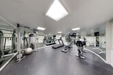 This fitness center is a well-appointed exercise facility designed to support a comprehensive workout routine. The space is defined by its clean, modern aesthetic, featuring crisp white walls and durable speckled rubber flooring that provides both safety and comfort underfoot. A striking diamond-shaped skylight serves as a central architectural feature of the ceiling, flooding the room with natural light and contributing to an open, airy atmosphere. Additional recessed and panel lighting ensures the space is consistently well-lit throughout. Floor-to-ceiling mirrors line multiple walls, visually expanding the room and allowing users to monitor their form during exercise. The equipment selection is thorough and varied, including treadmills, elliptical machines, a stationary bike, a cable strength training machine, and a stability ball, catering to both cardiovascular and resistance training needs. A wall-mounted flat-screen television is positioned for easy viewing during workouts. Horizontal window blinds along one wall offer adjustable natural light and privacy. The facility is accessible via a glass-paneled entry door, with hand sanitizer dispensers mounted near the entrance for hygiene convenience. Overall, the space presents a functional, professionally maintained environment suited for a full range of fitness activities.