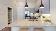 Located at 700 Corporate Center Drive in Raleigh, NC, this single-floor residence combines contemporary design with thoughtful functionality throughout. The kitchen is a standout feature, outfitted with sleek gray shaker-style cabinetry, stainless steel appliances including a French door refrigerator, electric range, and built-in microwave. A generous granite island with an undermount sink and matte black pendant lighting serves as both a prep and gathering space, while wood-look flooring anchors the room with warmth. The primary bedroom offers an elevated sense of calm with tall ceilings, soft neutral tones, and direct access to a well-designed en-suite bathroom. The bathroom features a dual-sink vanity with gray cabinetry, granite countertops, matte black fixtures, and wood-look flooring. A second bedroom provides ample natural light through large windows that frame outdoor greenery, complemented by neutral walls and hardwood-style floors. The private balcony extends the living space outdoors, offering a serene spot with views of landscaped surroundings and mature trees. Throughout the residence, a cohesive palette of soft grays, warm wood tones, and matte black accents creates a polished, modern aesthetic.
