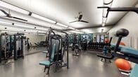 Located at 7510 Burgoyne in Houston, TX, this fitness center offers a comprehensive workout environment across a single, open floor. The space is thoughtfully organized into distinct exercise zones, each designed to support a full range of fitness activities.

The strength training area is well-appointed with multiple cable and resistance machines, free-weight benches, and an extensive dumbbell rack. Floor-to-ceiling mirrors line the walls, amplifying the sense of space and allowing users to monitor their form throughout their workouts. Ceiling fans and overhead fluorescent lighting ensure the area remains bright and comfortable.

A dedicated cardio zone features a row of Life Fitness treadmills and additional cardiovascular machines, accompanied by teal accent walls and framed motivational artwork that contribute to an energetic atmosphere. Natural light filters through large windows and glass double doors, providing views of the surrounding greenery.

A separate functional fitness area rounds out the space, equipped with stability balls in multiple sizes, resistance benches, and additional free weights. This zone is well-suited for stretching, core training, and accessory exercises. The durable rubber-tile flooring throughout the facility provides traction and impact absorption. Overall, the facility delivers a clean, organized, and fully functional environment for a wide variety of fitness needs.
