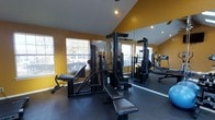 This single-level fitness space is designed to support a well-rounded workout routine within a compact, efficiently arranged layout. The room features vaulted ceilings that add vertical dimension and an open, airy feel despite the modest footprint. Bold yellow walls create an energetic atmosphere, complementing the dark rubber flooring that provides both durability and safety underfoot.

A large wall-to-wall mirror runs along one side of the room, visually expanding the space while allowing users to monitor their form during exercise. Natural light filters in through multiple windows on the opposite wall, offering views of surrounding greenery and contributing to a bright, welcoming environment.

The equipment layout includes a multi-station strength machine with a variety of attachment points for targeting different muscle groups, along with a weight bench for free weight exercises. A treadmill is visible in the mirrored reflection, indicating the room also accommodates cardiovascular training. Recessed lighting supplements natural light, ensuring the space remains well-lit at all hours. Overall, the room provides a focused and functional environment for both strength training and cardio workouts.