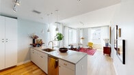 This well-appointed two-bedroom, two-bathroom residence in New Orleans offers a thoughtfully designed open-concept layout with a bright, modern aesthetic. Light wood flooring flows throughout the main living areas, complementing the crisp white walls and abundant natural light streaming through large windows.

The kitchen serves as the heart of the home, anchored by a sleek island with pendant lighting overhead, stainless steel appliances, and white cabinetry with brass hardware. It opens seamlessly to the dining area and spacious living room, where floor-to-ceiling windows frame sweeping views of the New Orleans skyline — filling the space with natural light and urban energy.

The primary bedroom is a generous retreat, featuring a bold upholstered accent wall in warm terracotta tones, large windows with city views, and light wood flooring. The second bedroom is equally inviting, with a ceiling fan, city views, and a soft, airy palette. Both bedrooms are served by well-organized walk-in closets with built-in shelving and hanging systems.

The two bathrooms are finished in a clean, contemporary style, with one featuring a slate-blue vanity, a gold pendant light, and a large framed mirror. The hallway connecting the rooms is bright and welcoming, accented with decorative touches that reflect the residence's curated character.