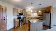This single-floor space showcases a thoughtfully designed kitchen that serves as its centerpiece. The kitchen features warm-toned wood cabinetry that lines the walls, providing ample storage alongside open shelving that adds a functional, airy quality to the layout. Stainless steel appliances, including a range with oven, over-the-range microwave, and refrigerator, lend a modern, polished look to the space.

A central island with a granite-style countertop anchors the kitchen, offering additional workspace and seating potential. Two pendant lights hang above the island, providing focused task lighting that complements the flush-mount ceiling fixture overhead. The sink area, positioned along a dedicated countertop run, includes under-cabinet lighting for added practicality.

The hardwood-style flooring in a warm honey tone runs throughout, tying the space together cohesively. An open shelving unit near a doorway provides additional storage or display space. The overall layout is efficient and open, with a neutral color palette of whites and warm wood tones creating a bright, welcoming atmosphere suited for everyday living.