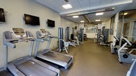 This fitness center is designed to support a well-rounded exercise routine, offering a thoughtfully arranged layout across a single-floor space. The room is outfitted with a row of treadmills and cardio machines along one side, complemented by multiple cable and weight-resistance machines positioned throughout the floor. Wall-mounted flat-screen televisions are installed near the cardio area, allowing users to stay entertained during their workouts. The flooring consists of durable rubber matting, providing both safety and comfort underfoot. Exposed ductwork runs along the ceiling, lending the space an industrial aesthetic, while a drop-ceiling section with recessed and panel lighting ensures the room is well-lit throughout. Windows along the far wall allow natural light to filter in and offer a view of the outdoors, enhancing the overall atmosphere. The neutral-toned walls and open arrangement of equipment create an uncluttered, functional environment. The space appears suitable for both cardio training and strength conditioning, making it a versatile facility for users of varying fitness levels and goals.