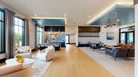 Located at Ascend Village in Winter Garden, Florida, this thoughtfully designed single-floor community clubhouse offers a generous and versatile gathering space. The open-plan main lounge is defined by large floor-to-ceiling windows that flood the interior with natural light and frame views of lush tropical landscaping. Warm golden tile flooring anchors the space, complemented by a striking teal accent ceiling with integrated cove lighting that adds a bold architectural focal point.

The lounge is divided into distinct seating areas—one features plush curved armchairs arranged around a low coffee table, while another offers a more formal arrangement with leather lounge chairs and a bar-height counter beneath a dramatic patterned backsplash, pendant lighting, and a mounted display screen. A separate carpeted lounge zone provides additional collaborative seating with a modern chandelier overhead.

Three private offices serve as quiet workspaces, each thoughtfully appointed. One functions as a conference room with a large dark wood table, gold-accented chairs, and tall windows overlooking the outdoors. Individual workstations feature built-in white desks set against textured wood-panel accent walls. A hallway with polished tile connects the spaces, leading to exterior access points, a closet, and utility areas, completing this well-appointed community hub.