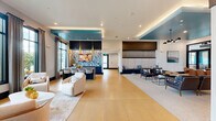 Located at Ascend Village in Winter Garden, Florida, this thoughtfully designed single-floor community clubhouse offers a generous and versatile gathering space. The open-plan main lounge is defined by large floor-to-ceiling windows that flood the interior with natural light and frame views of lush tropical landscaping. Warm golden tile flooring anchors the space, complemented by a striking teal accent ceiling with integrated cove lighting that adds a bold architectural focal point.

The lounge is divided into distinct seating areas—one features plush curved armchairs arranged around a low coffee table, while another offers a more formal arrangement with leather lounge chairs and a bar-height counter beneath a dramatic patterned backsplash, pendant lighting, and a mounted display screen. A separate carpeted lounge zone provides additional collaborative seating with a modern chandelier overhead.

Three private offices serve as quiet workspaces, each thoughtfully appointed. One functions as a conference room with a large dark wood table, gold-accented chairs, and tall windows overlooking the outdoors. Individual workstations feature built-in white desks set against textured wood-panel accent walls. A hallway with polished tile connects the spaces, leading to exterior access points, a closet, and utility areas, completing this well-appointed community hub.