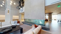 Located on West Pico Boulevard in Los Angeles, this contemporary amenity floor delivers a striking blend of modern architecture and refined interior design. The centerpiece of the space is a dramatic double-height lounge anchored by a monumental concrete wall and a wide, glass-enclosed fireplace, creating a bold focal point visible from multiple seating areas. Oversized sculptural pendant lights hang from the soaring ceiling, lending artistic character to the voluminous room. Plush, thoughtfully arranged seating groupings invite gathering and relaxation throughout. A fully equipped kitchen features an expansive marble-topped island with bar seating, light wood cabinetry extending to the ceiling, a veined stone backsplash, and stainless steel appliances including a built-in wine refrigerator. Adjacent lounge and co-working areas offer comfortable seating alongside natural wood accents and vibrant geometric-patterned rugs. A dedicated media lounge with a slatted wood ceiling and large-screen display provides an intimate entertainment setting. Floor-to-ceiling windows frame panoramic views of the surrounding Los Angeles cityscape, filling the space with abundant natural light. The overall aesthetic is cohesive and upscale, balancing warm natural materials with cool stone and concrete finishes throughout.