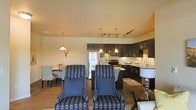This single-floor residence offers a thoughtfully designed open-concept layout that seamlessly connects the living, dining, and kitchen areas into one cohesive and functional living space. The living area features patterned accent chairs in a bold blue and white design, complemented by warm wood-tone flooring that extends throughout. A decorative wall-mounted stag head adds a distinctive, characterful touch to the space. The dining area is positioned adjacent to the living space, furnished with a classic table and seating, ideal for everyday meals or entertaining. The kitchen is a standout feature, showcasing rich dark cabinetry that extends to the upper walls, stainless steel appliances including a refrigerator and microwave, and a central island or peninsula with pendant lighting overhead. A track lighting system provides additional illumination, ensuring the kitchen remains bright and functional. The countertops offer ample prep space, and the layout promotes an efficient workflow. Neutral-toned walls and consistent flooring throughout the space create a unified, welcoming ambiance. The open design allows for excellent sightlines across all areas, making the space feel expansive and connected while maintaining distinct zones for living, dining, and cooking.