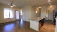 This single-story space is designed with an open-concept layout that seamlessly connects the kitchen and living areas, creating a functional and cohesive flow. The kitchen is a standout feature, anchored by a peninsula-style island with a granite-look countertop and a double-basin stainless steel sink. Dark cabinetry lines the kitchen and is complemented by a stainless steel dishwasher, while a pendant light fixture suspended above the island provides focused task lighting and a touch of style.

The flooring throughout is a rich, dark hardwood-style material that adds warmth and visual continuity across the space. The living area extends beyond the kitchen, benefiting from natural light streaming through multiple windows fitted with horizontal blinds. A ceiling fan is visible in the living space, adding both practicality and a casual finish to the area.

The neutral wall tones in warm beige create a versatile backdrop throughout, while vaulted or slightly elevated ceilings in the living zone add a sense of openness. Additional doorways visible in the background suggest further rooms beyond the main living area, contributing to the overall functional layout of the space.