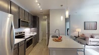 This single-floor unit is designed with a clean, modern aesthetic and a neutral color palette that creates a cohesive, open feel throughout. The kitchen serves as a central highlight, featuring dark espresso cabinetry with brushed hardware, white quartz countertops, a subway tile backsplash, and a full suite of stainless steel appliances including a refrigerator, range, and over-the-range microwave. A peninsula with an undermount sink and pendant lighting overhead provides casual seating and separates the kitchen from the living area, where light wood-style flooring continues underfoot. The living room offers natural light through a large window with views of the surrounding greenery, complementing the warm, understated atmosphere. Two bedrooms feature plush carpeting, ceiling fans, and windows that bring in additional natural light. The primary bathroom is finished with a wide vanity, quartz countertop, undermount sink, tile flooring, and a glass-enclosed shower. A secondary bathroom offers a tub-shower combination with white subway tile surround and tile flooring. Throughout the unit, consistent neutral tones and modern fixtures contribute to a polished, comfortable living environment.