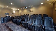 This private theatre is designed to deliver a true cinematic experience in a refined, comfortable setting. The room is arranged with multiple rows of plush, dark upholstered reclining seats, each equipped with built-in cup holders — ensuring every seat offers both comfort and convenience. The seating is tiered to provide clear sightlines throughout the room, accommodating a generous number of guests.

A ceiling-mounted projector serves as the centerpiece of the audiovisual setup, positioned to deliver a crisp, large-format image to the front screen. Recessed lighting is embedded into the ceiling, providing soft, dimmable illumination that enhances the theatre atmosphere without detracting from the on-screen experience. Crown molding runs along the upper walls, adding a touch of architectural elegance to the space.

The walls are finished in warm, neutral tones that complement the dark seating and patterned carpeted flooring, which features a decorative design that adds visual interest underfoot. A framed artwork piece adorns the rear wall, contributing to the welcoming ambiance. The overall design balances functionality with sophistication, making this theatre an ideal venue for film screenings, presentations, or private events.