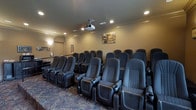 This private theatre is designed to deliver a true cinematic experience in a refined, comfortable setting. The room is arranged with multiple rows of plush, dark upholstered reclining seats, each equipped with built-in cup holders — ensuring every seat offers both comfort and convenience. The seating is tiered to provide clear sightlines throughout the room, accommodating a generous number of guests.

A ceiling-mounted projector serves as the centerpiece of the audiovisual setup, positioned to deliver a crisp, large-format image to the front screen. Recessed lighting is embedded into the ceiling, providing soft, dimmable illumination that enhances the theatre atmosphere without detracting from the on-screen experience. Crown molding runs along the upper walls, adding a touch of architectural elegance to the space.

The walls are finished in warm, neutral tones that complement the dark seating and patterned carpeted flooring, which features a decorative design that adds visual interest underfoot. A framed artwork piece adorns the rear wall, contributing to the welcoming ambiance. The overall design balances functionality with sophistication, making this theatre an ideal venue for film screenings, presentations, or private events.