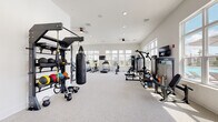 This single-story fitness center is designed to accommodate a wide range of workout styles within a bright, open layout. The space is divided into distinct functional zones, each outfitted with purpose-specific equipment. The strength and functional training area features a wall-mounted multi-station rig complete with a heavy bag, TRX suspension trainers, kettlebells, and an assortment of medicine balls. Adjacent to this, a dedicated cable and resistance machine zone provides additional strength training options.

The cardio section is well-appointed with treadmills, an elliptical, a rowing machine, and a stationary bike, positioned near large windows that invite ample natural light and offer views of the surrounding community and outdoor pool area. A separate cycling studio features a full wall of floor-to-ceiling mirrors, spin bikes, and a wall-mounted television, creating an immersive group or solo training environment.

Throughout the space, neutral-toned carpet flooring, crisp white walls, recessed lighting, and ceiling fans contribute to a clean, modern ambiance. Multiple wall-mounted televisions are distributed across the facility. The expansive windows on multiple walls ensure the space remains bright and visually connected to the outdoors.